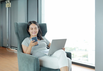 Young beautiful asian woman using smartphone and laptop computer working online at home and feel relaxing on armchair. People during the introvert time at home from the epidemic in thailand.