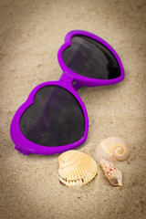 Fototapeta na wymiar Purple sunglasses in shape of heart with shells on sand