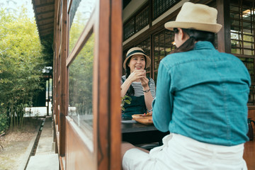 shot from outdoor through window and door with two asian girl friends sitting on wooden floor in ryokan and smiling drinking tea. beautiful women laughing enjoy tea ceremony by sunny spring garden