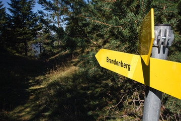 Yellow hiking signs on a pole pointing towards a hiking trail to Brandenberg, Austria in a pine forest.
