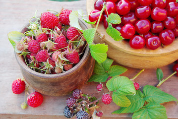 Two wooden bowls with raspberries and cherries on a wooden board.