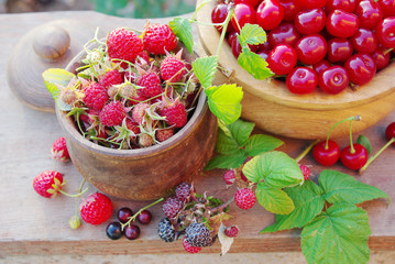 Two wooden bowls with raspberries and cherries on a wooden board.