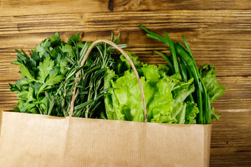 Paper bag with green onion, rosemary, lettuce leaves and parsley on wooden table. Top view. Healthy food and grocery shopping concept
