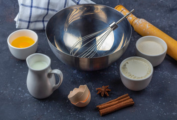 Baking background. Empty metal bowl among ingredients and utensils for cooking cake (flour, egg, milk, sugar, rolling pin, towel) on dark table. Food concept. Top view, flat lay, copy space for text