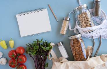 list and notebook for zero waste lifestyle .cotton net bag with fresh vegetables and sustainable glass jar on blue background flat lay.plastic free for groceries product shopping and delivery.