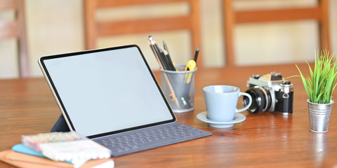Photo of white blank screen computer tablet with keyboard case that putting on wooden working desk and surrounded by coffee cup, camera, potted plant, stationary in cup and stack of notebook.