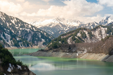 Fototapeta na wymiar Beautiful nature background and wallpaper of green reservoir lake at Kurobe dam surrounded by snow mountain of Japan Alps under blue sky with cloud and sun light. Top tourist destination in Nagano
