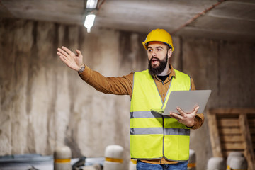 Young serious attractive contractor standing with laptop in hands and giving orders to workers.