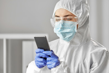 health safety, medicine and pandemic concept - close up of female doctor or scientist in protective wear, medical mask, gloves and goggles for protection from virus disease with smartphone