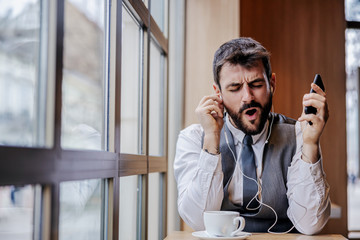 Young cheerful bearded businessman sitting in cafeteria, listening music over smart phone and singing.