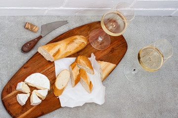 Two glasses of white wine, camemebert, cheese knife and baguette on wooden board.