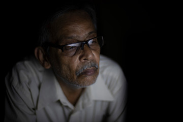 An Indian old man is watching laptop screen while video chatting leaning on his bed in the darkness. Indian lifestyle, home isolation and lock down
