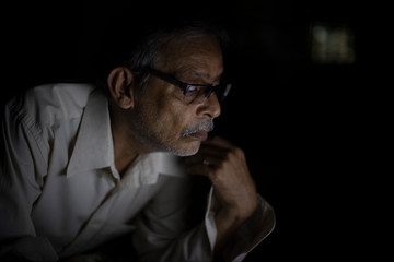 Fototapeta premium An Indian old man is watching laptop screen while video chatting leaning on his bed in the darkness. Indian lifestyle, home isolation and lock down