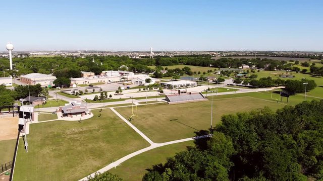 Moving diagonally towards football field, "CITY OF HUTTO" at the top of the scoreboard sign. Large portion of park visible in back ground including pavilion, tennis,basketball, & volleyball courts.