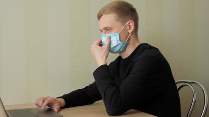 Man in a protective medical mask works at home with laptop. The guy sits on chair at the table, looks at the laptop screen and straightens the mask. Quarantine, self-isolation