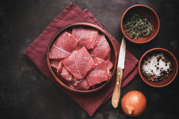 Fresh raw pieces of beef meat in a bowl with spices, herbs and onions on a dark background. Top view, flat lay, copy space.