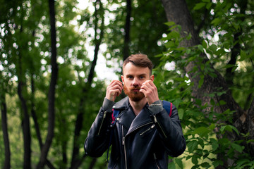 Stylish guy straightens his orange mustache while standing near green trees in a black leather jacket.