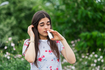 Fototapeta premium Pollen allergy concept. Young woman using nose spray for her pollen and grass allergies. Flowering trees in background. Spring Seasonal allergies and health problems.