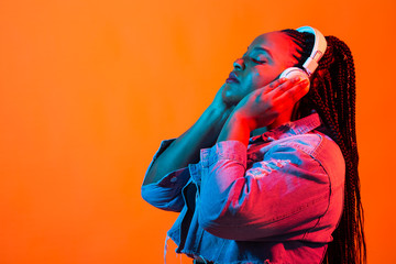 Close up beauty fashion portrait of an attractive young african woman wearing dreadlocks standing over orange background, listening to music with headphones. Neon light and copy space