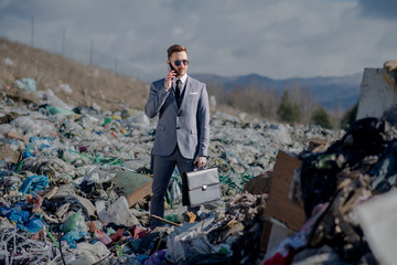 Businessman with smartphone on landfill, consumerism versus pollution concept.