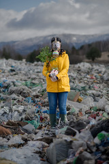 Fototapeta na wymiar Woman with gas mask holding green plant on landfill, environmental concept.