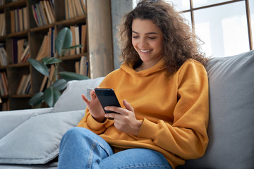 Happy millennial hispanic teen girl checking social media holding smartphone at home. Smiling young latin woman using mobile phone app playing game, shopping online, ordering delivery relax on sofa.