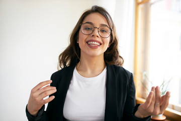 Indoor shot of friendly confident millennial female vlogger in stylish round eyeglasses speaking at...