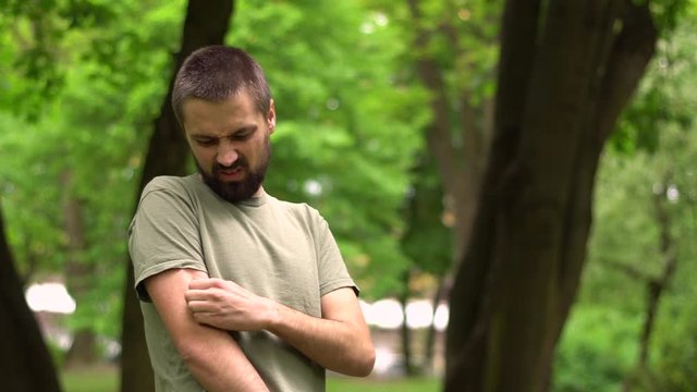 A caucasian young bearded man scratches his hand because of an allergy in the park.
