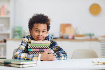 Portrait of cute African-American boy holding stack of books while sitting at desk at home and...