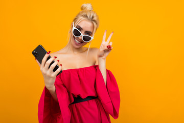 charming lady in a red dress in sunglasses on her face holds a phone and shows a welcome sign on a yellow background with copy space