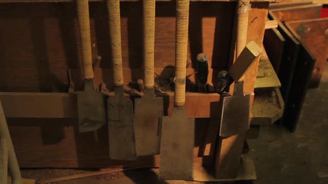 Cord wrapped handles of antique cutting tools and saws in Japanese artisan carpentry shop. Traditional craftsmanship implements. Old hand tools hang on work bench with golden light. Zoom out.