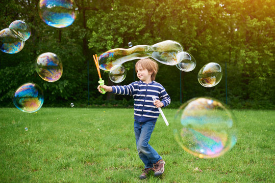 Outdoors portrait of cute preschool boy blowing soap bubbles on a green lawn at the playground.