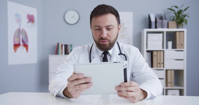 Medium plan of handsome smart therapist in office in white medical gown with statoscope talking to client using his tablet. The doctor consultig online. Сoncept of pandemic, modern medicine.