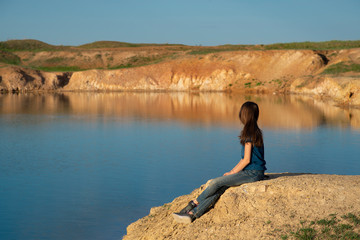 Fototapeta na wymiar A girl sits on the shore of a blue lake on a summer sunny day.