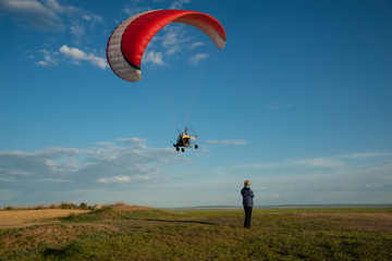 Motorized paraglider flies in the blue sky. Woman in a field looks from below.