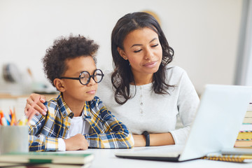 Portrait of cute African boy wearing big glasses while using laptop with mom, homeschooling and remote education concept