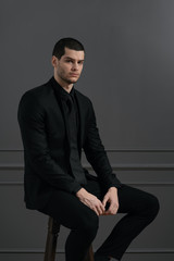 Young handsome businessman in black shirt and black suit. Confident young businessman posing on gray background sitting on chair