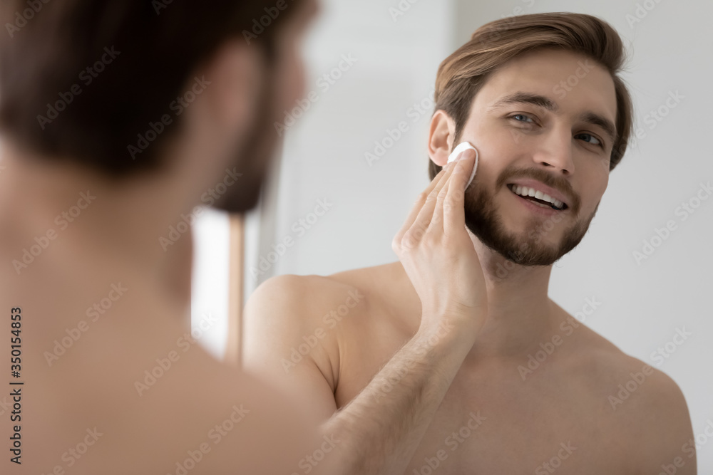 Wall mural Smiling young Caucasian metrosexual man look in mirror in bathroom apply facial toner or lotion with cotton pad, happy millennial handsome male do morning beauty face procedures, skincare concept