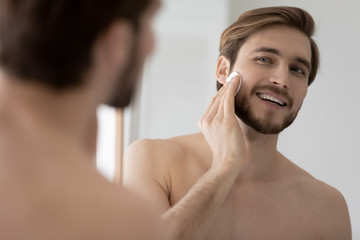 Smiling young Caucasian metrosexual man look in mirror in bathroom apply facial toner or lotion...