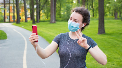 Young woman in medical mask using mobile phone for video call,showing peace sign with two fingers during walking in park outdoor. Selfie. Using wireless Internet connection.Active life during covid