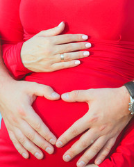 Married couple hold hands in the form of a heart on the stomach of a pregnant girl.Pregnant woman
in a red and man in a white shirt on the lake.The concept of a happy marriage and love. Close-up