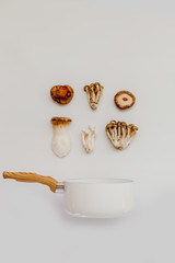 A variety of wild mushrooms and a white saucepan with wooden handle on a pastel white background in a flat lay composition