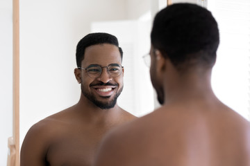 Fototapeta na wymiar Smiling naked young african American man in glasses look in mirror in bathroom after shower, happy handsome millennial biracial male in spectacles show white healthy teeth in bath, hygiene concept