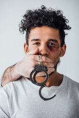 Male hand holding handcuffs and covering mouth to african american man with bruise isolated on grey