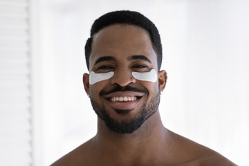 Headshot portrait pf smiling young african American man use apply under eye anti-wrinkle patches in bath, happy biracial male do morning beauty procedures for healthy glowing skin, skincare concept