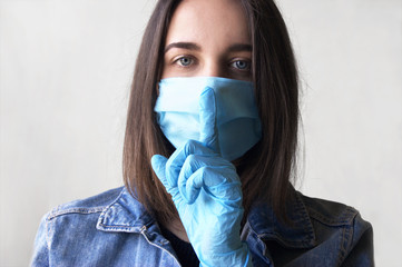 A young woman wears a medical mask and shows a gesture of silence with a finger on her lips.