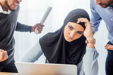 Selective focus of upset muslim businesswoman looking at camera near quarreling colleagues in office