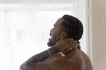 Happy young sexy african American man stand under water flow enjoy taking daily morning shower in...