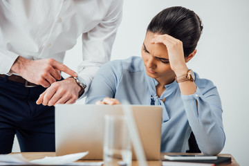 Selective focus of businessman pointing at wristwatch near sad mexican colleague using laptop in...