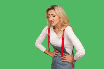 Stomach pain. Portrait of sick adult woman in denim overalls suffering abdominal cramps, spasms and aching belly from indigestion, gastrointestinal disorder. studio shot isolated on green background